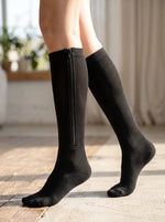 A person wearing black knee-high socks with a visible side zipper, shown standing on a light stone floor in a softly lit indoor setting. 